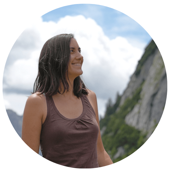A girl with brown hair with a smile on her face she facing away from the camera, natural setting in the background.